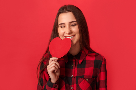 Valentines Day. Young girl standing isolated on red biting valentine card laughing cheerful close-upの写真素材