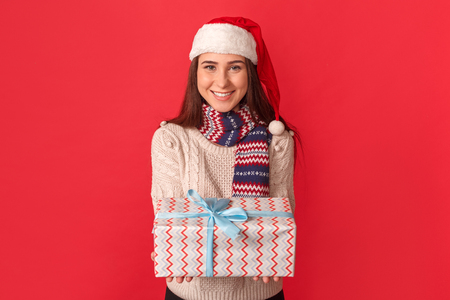 Celebration Concept. Young woman in scarf and santa hat standing isolated on red giving present to camera smiling cheerfulの写真素材