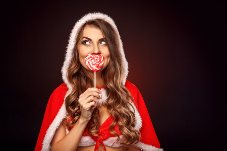 Christmas and New Year. Woman in santa costume with hood standing isolated on black covering mouth with lollipop looking aside playfulの写真素材