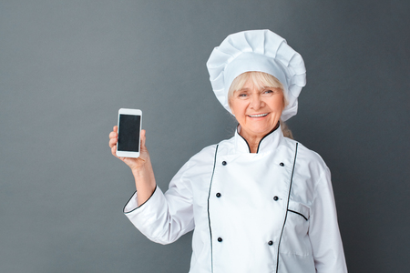 Senior woman chef studio standing isolated on gray with smartphone showing screen looking camera cheerfulの写真素材