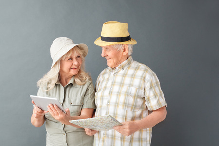 Senior tourists in beach hats studio standing isolated on gray holding map using digital tablet looking at each other playfulの写真素材