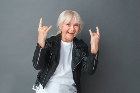 Senior woman in leather jacket studio standing isolated on gray showing horns with two hands looking camera cheerfulの写真素材