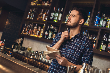 Young bartender standing at bar counter holding notebook thinkingの写真素材