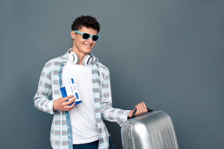Man in sunglasses isolated on gray wall tourism concept standing with suitcase holding passport and ticket cheerfulの写真素材