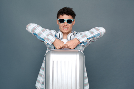 Man in sunglasses isolated on gray wall tourism concept standing lifting up suitcase grimaceの写真素材