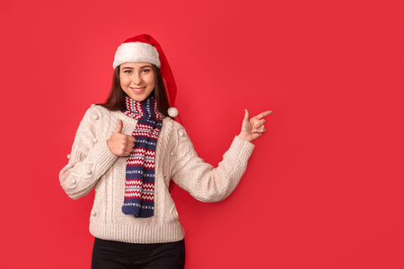 Celebration Concept. Young woman in scarf and santa hat standing isolated on red thumb up pointing aside smiling positiveの写真素材