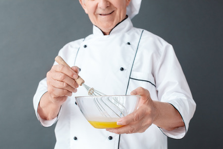 Senior chef studio standing isolated on gray whisking eggs cheerful close-upの写真素材