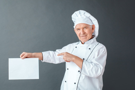 Senior chef studio standing isolated on gray pointing at paper looking camera playfulの写真素材