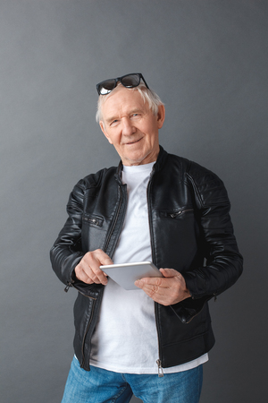 Senior man in leather jacket standing isolated on gray playing game on digital tablet looking camera cheerfulの写真素材