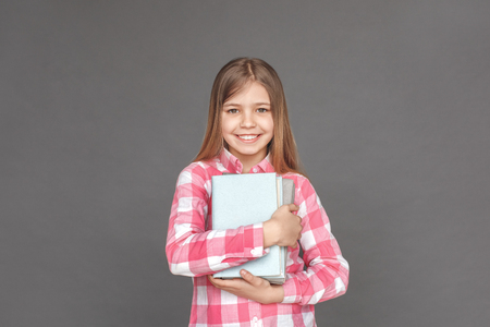 Good schoolgirl. Girl standing isolated on grey with pile of books close smiling joyfulの写真素材