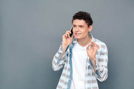 Man isolated on gray wall free style standing talking on smartphone showing ok gesture friendlyの写真素材