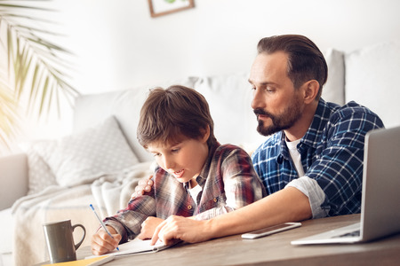 Father and son at home sitting at table dad showing boy how to do assignmentの写真素材