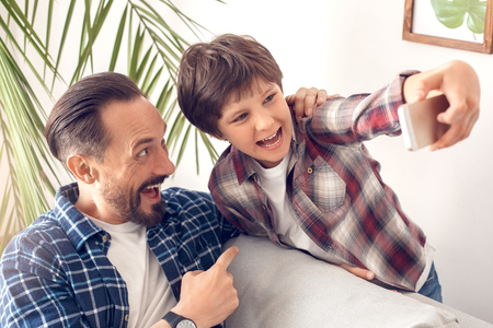Father pointing at little son shouting playful taking selfie on smartphone at homeの写真素材