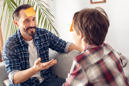 Father and little son at home sitting on sofa playing forehead detective talking joyfulの写真素材