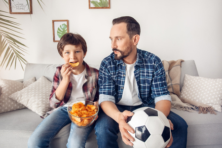 Father and little son at home sitting on sofa dad with ball looking at boy holding potato chip watching football smiling excitedの写真素材