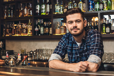 Young bartender leaning on bar counter smiling restrainedの写真素材