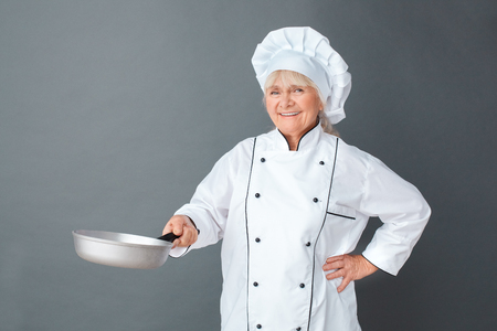 Senior woman chef studio standing isolated on gray with frying pan cooking looking camera joyfulの写真素材