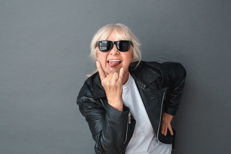 Senior woman in leather jacket and sunglasses studio standing isolated on gray showing tongue through the horns sign coolの写真素材