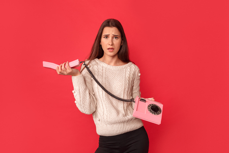 Freestyle. Young woman standing isolated on red with stationary phone handset looking camera concernedの写真素材