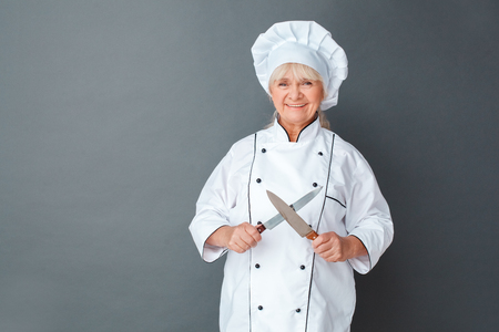 Senior woman chef studio standing isolated on gray sharpening knives looking camera cheerfulの写真素材
