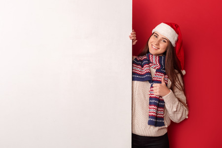 Freestyle. Young woman wearing scarf and santa hat standing isolated on red with white board thumb up looking camera cheerfulの写真素材