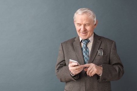 Senior man studio standing isolated on gray typing message on smartphone smiling cheerfulの写真素材