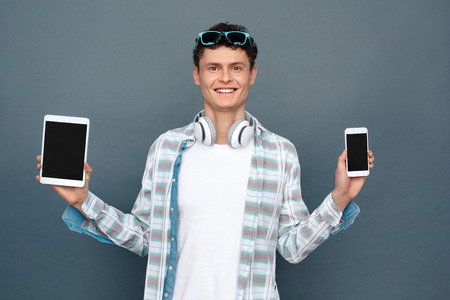 Man isolated on gray wall tourism concept standing holding smartphone and digital tablet looking camera happyの写真素材