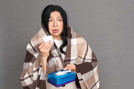 Freestyle. Mature lady in plaid blanket standing isolated on grey with tissue box having runny noseの写真素材