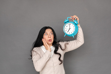 Freestyle. Mature lady standing isolated on grey with alarm clock up looking at time shockedの写真素材