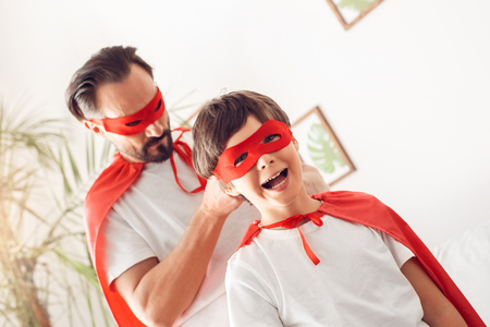 Father and son in superheroe costumes at home standing man tying knot on mask of boy smiling cheerfulの写真素材