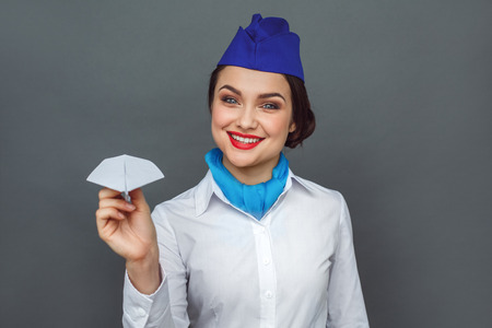 Professional Occupation. Stewardess standing isolated on grey with paper plane smiling cheerful close-upの写真素材