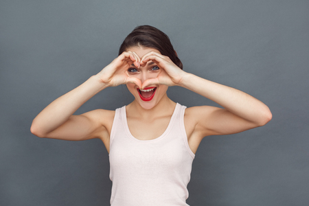 Freestyle. Woman standing isolated on grey looking through heart shape laughing playfulの写真素材