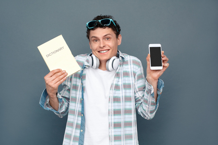 Man isolated on gray wall tourism concept standing holding dictionary and smartphone choosing option confusedの写真素材