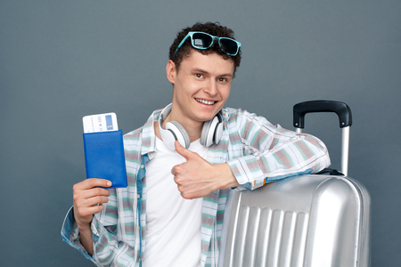 Man isolated on gray wall tourism concept standing leaning on suitcase holding passport with ticket thumb up looking camera positiveの写真素材