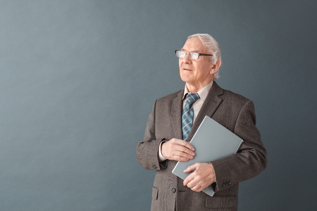 Senior man teacher wearing glasses studio standing isolated on gray holding book looking aside thoughtfulの写真素材