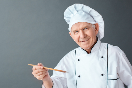Senior chef studio standing isolated on gray holding spatula looking camera positive close-upの写真素材