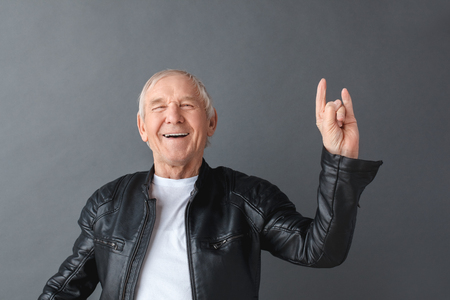 Senior man in leather jacket standing isolated on gray showing horns gesture laughing joyful close-upの写真素材