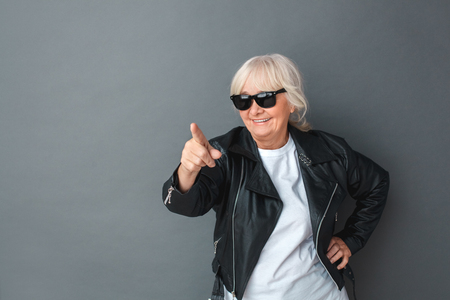 Senior woman in leather jacket and sunglasses studio standing isolated on gray pointing camera playfulの写真素材