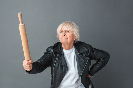 Senior woman in leather jacket studio standing isolated on gray with rolling pin threatening someone angryの写真素材