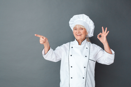 Senior woman chef studio standing isolated on gray showing ok pointing at space asdie happyの写真素材
