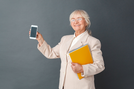 Senior woman in fromal suit and glasses studio standing isolated on gray holding journal howing smartphone screen smiling happyの写真素材