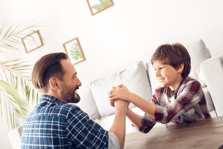 Father and little son at home sitting at table arm wrestling boy holding dads hand smiling cheerfulの写真素材
