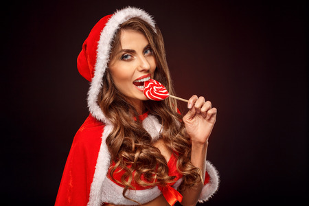 Christmas and New Year. Woman in santa costume with hood standing isolated on black biting lollipop smiling playful close-upの写真素材