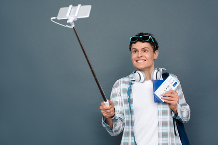 Man isolated on gray wall tourism concept standing holding monopod taking selfies on phone showing passport and ticket looking camera smilingの写真素材