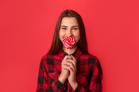 Freestyle. Young girl standing isolated on red covering mouth with heart shaped lollipop laughing cheerfulの写真素材