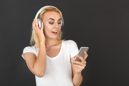Freestyle. Woman in headphones standing isolated on black wall choosing song on smartphone listening music surprisedの写真素材