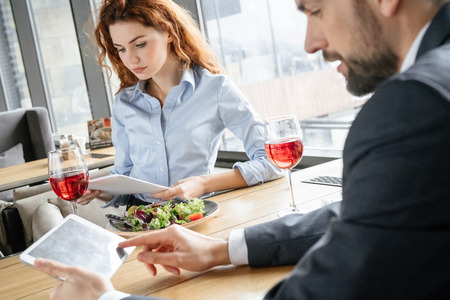 Businesspeople having business lunch at restaurant sitting eating salad drinking wine man using digital tablet while woman reading documentsの写真素材