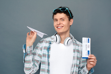 Man isolated on gray wall tourism concept standing holding origami plane and ticket looking camera cheerfulの写真素材