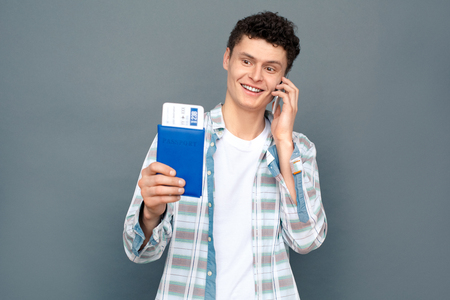 Man isolated on gray wall tourism concept standing holding passport with ticket talking on phone excited cheerfulの写真素材