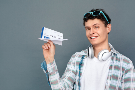 Man isolated on gray wall tourism concept standing holding origami plane with ticket looking camera joyfulの写真素材